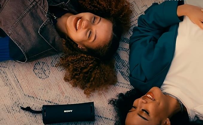 Two friends lying on a rug, smiling, with a portable Sony speaker beside them, enjoying music together